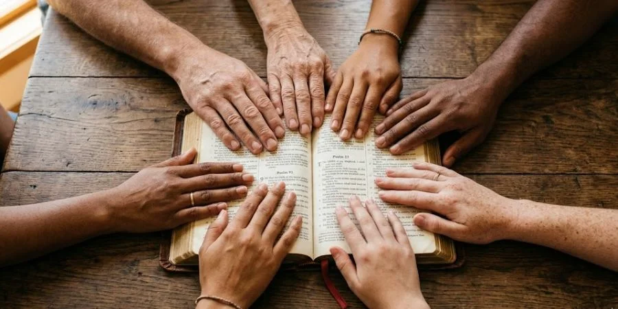 Diverse hands gathered over an open Bible in warm light, illustrating Bible verses about identity as a chosen and holy community.